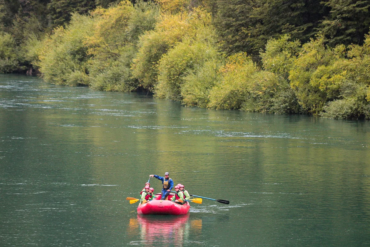 Family paddling a raft on a calm stretch of green water, surrounded by native forest and cloud-capped mountains